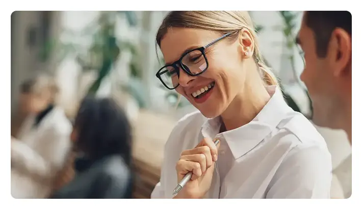 A professional woman with glasses is smiling while holding a pen and speaking with a colleague in an office.